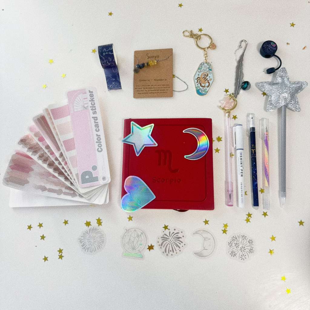 Collection of stationery items including a red notebook, stickers, and pens on a white background with star decorations.